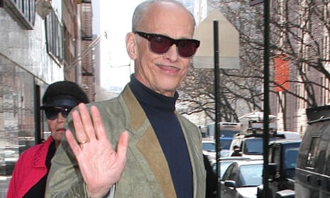 John Waters in dark glasses on a street, holding up a hand