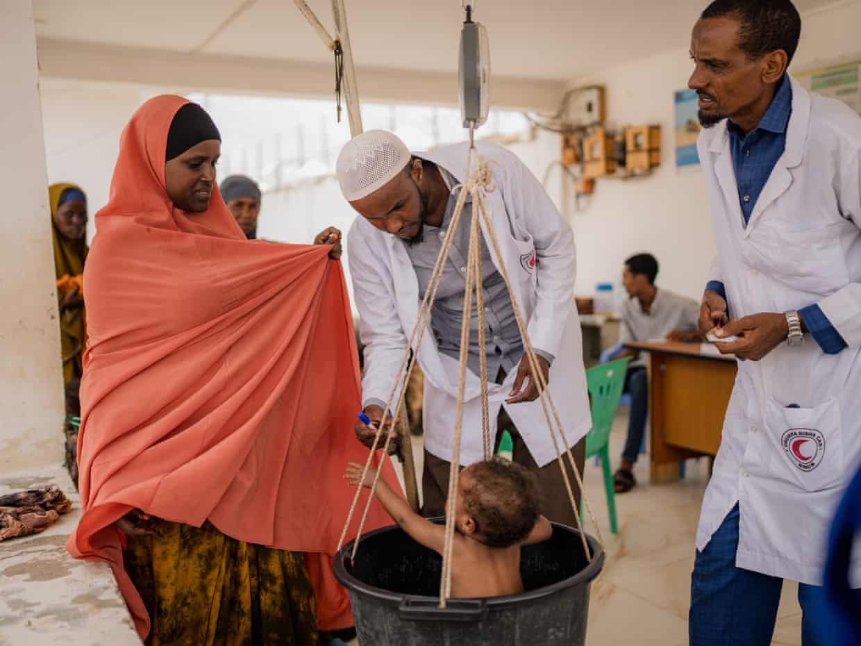 A woman has her child weighed at a health centre, watched by doctors in white coats