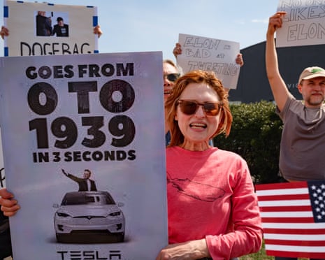 a woman with a placard showing Elon Musk leaning out of the car giving a fascist-style salute, the placard states 'Goes from0-1939 in 3 seconds
