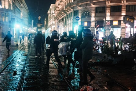 Police with riot shields and helmets clash with people in the square with luxury shops in the background. It is dark and raining, with blue lights reflecting off the stone paving.