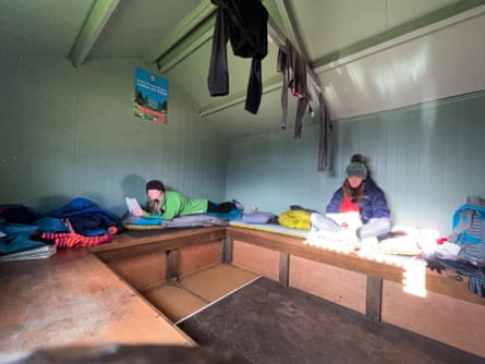 Phoebe and Ellie wearing warm clothes and sprawling on wooden benches in the bothy, with clothes hung up to dry on a rafter