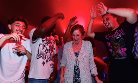 An older woman wearing a cardigan with three younger men in T-shirts raising their hands in the air.