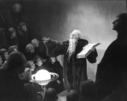 In Faust, a bearded elderly man gestures while holding an open book near a glowing orb