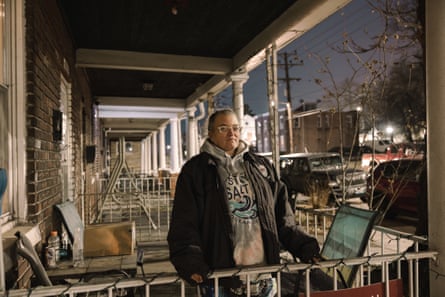 a wide portrait of a person in a gray hoodie and black jacket rests her hands on the railing of her porch with a row of homes visible behind her and power lines and lights in the background