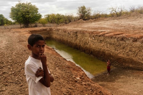 Two children stand next to a deep and long trench cut into rocky soil and partly full of greenish water
