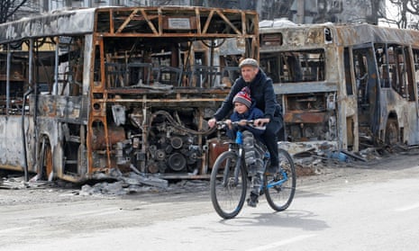A man and a child ride a bicycle past burnt out buses in Mariupol.