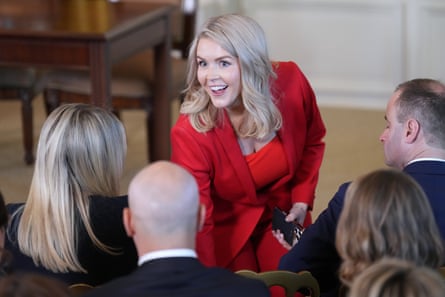 A blonde woman in a red suit smiles and greets people who are seated in a room
