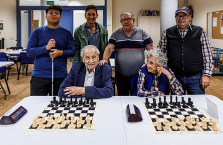 An elderly man and woman sit in front of chess boards, while their fellow club members stand behind.