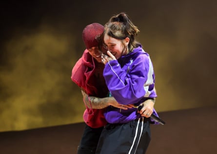 Billie Eilish looks overwhelmed on stage with Justin Bieber, who is hugging her. Both are smiling widely