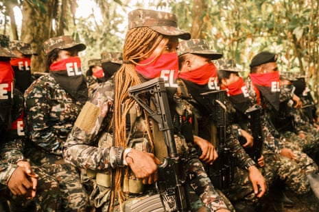 A group of fighters in combat gear with their noses and mouths covered in red and black bandanas standing with their rifles in a jungle clearing