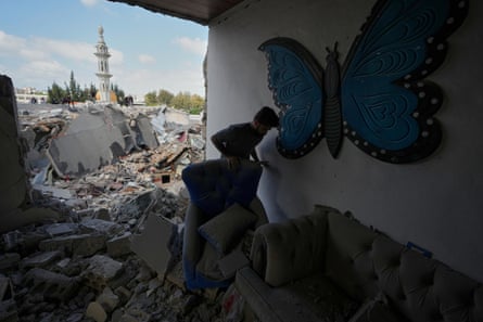 A man stands by an armchair in a pile of rubble in a room that is open to the elements. A tall spire is in the background among destroyed buildings. There is a large decorative blue butterfly on the wall of the room.