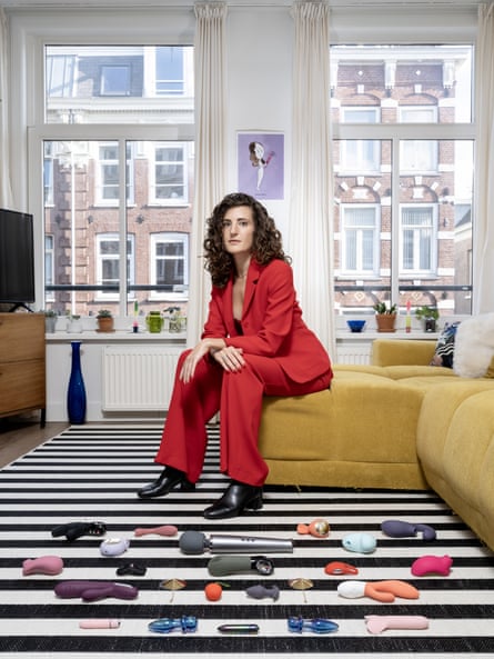 Portrait of Jessica sitting on a yellow sofa in front of two windows with her sex toys laid out on a black and white striped rug