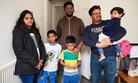 Akhil Jenny stands behind a young family with three small children, two boys and a girl; their father holds the girl in his arms and is smiling but the other two adults look serious. They are standing in front of a doorway inside a room with white-painted walls, a chipped wooden table and children’s clothing hanging up.