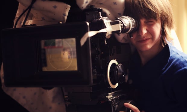 A young woman operating a 35mm film camera. There is a blanket thrown over the camera to dampen the sound of the film running through it so it doesn't interfere with the scene