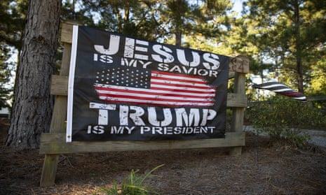 A flag that say "Jesus is my savior/Trump is my president" alongside an American flag tacked to a wooden fence amid trees along a rural road.