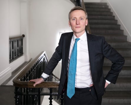 Torsten Bell stands on a stairway in the Department for Work and Pensions with a flight of dark-carpeted stairs behind him. He is leaning on a wooden banister on a landing and wears an open dark jacket, white shirt, and a slightly askew pale blue tie. He is in his early 40s with short fair hair.