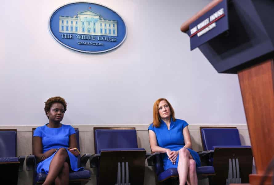 Two women wearing blue dresses sit in chairs below an emblem of the presidential seal.