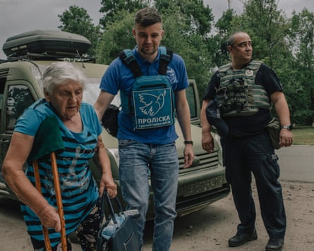 Elderly woman using a wooden crutch is helped to walk by two policemen in flak jackets