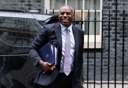 David Lammy arrives at Downing Street. He walks in front of black iron railings and holds folders of documents under his arms, and is smiling.