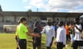 A man in a neon green jersey shaking hands on a football pitch with a man in a white shirt