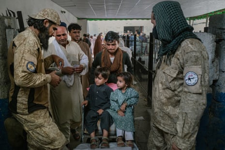 A man with two young children in a wheeled chair and an older man beside him passes between military personnel in a corridor; the older man is holding a paper document