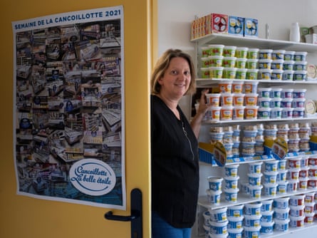 Julie Morin stands behind a yellow door and next to rows of cheese