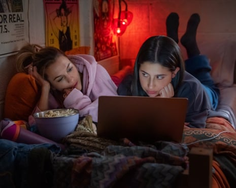 Two young women on their stomachs on a twin bed, looking at a laptop.