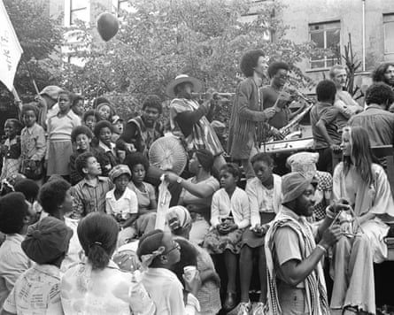 A black and white photo of a crowd sitting on a mound at Notting Hill Carnival, some playing instruments, others holding flags in 1974