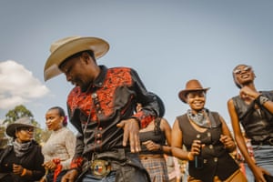 A man in a cowboy hat and black shirt with red detailing dances in front of people wearing western hats and neckerchiefs