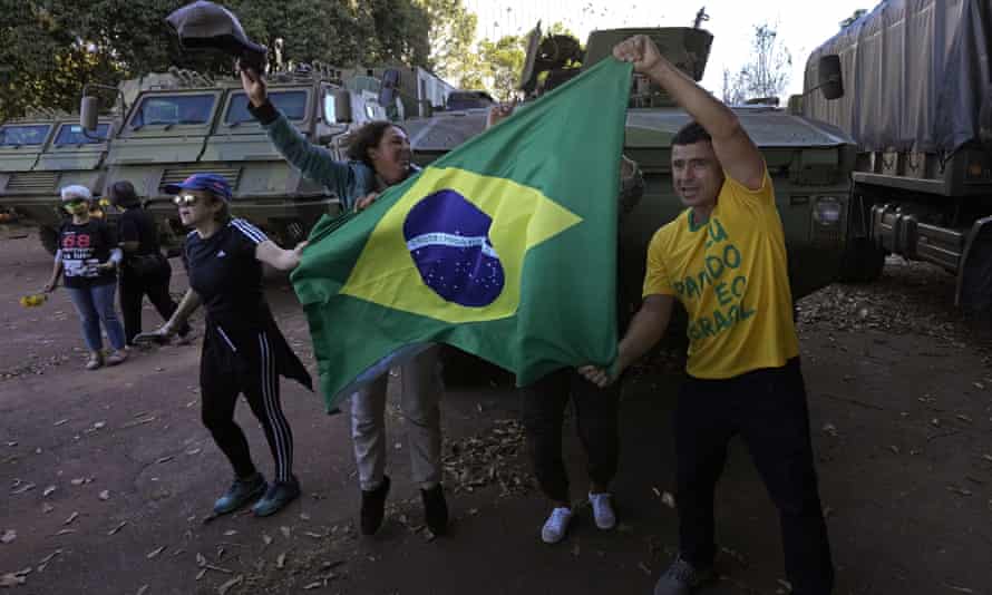 Apoiadores de Jair Bolsonaro comemoram com uma bandeira brasileira enquanto manifestantes seguram flores para dar aos soldados do comboio militar em Brasília