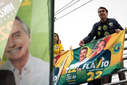 Nikolas Ferreira leans over a balcony behind an election banner which has a picture of Flavio Bolsonaro on it.