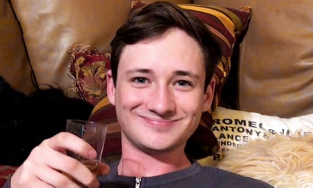 A young white man with brown hair, smiling as he leans back on a couch pillow, holding up a small glass.