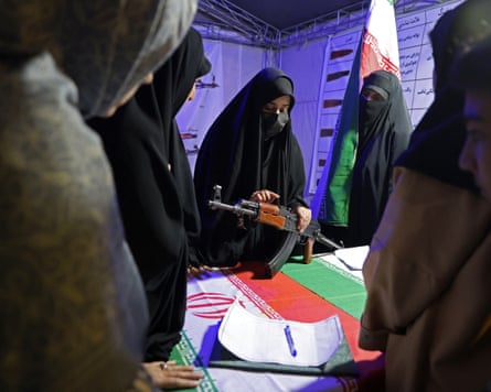 A woman in a burqa shows other women how to use a gun. There is an Iranian flag on the table.