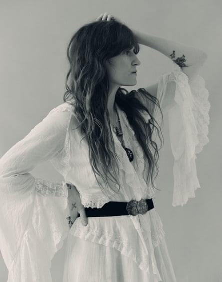 Florence Welch standing up looking to her left, with one hand on her head and the other on her waist, wearing a flowing white dress with a black belt
