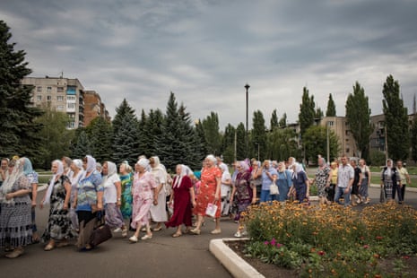 A group of people – mainly older women wearing headscarves and some men – walk in a line through a town gardens on an overcast day