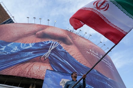 A man waves an Iranian flag under a billboard with graphic showing the strait of Hormuz and the sewn lips of Donald Trump in a square in downtown Tehran.