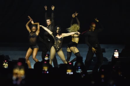 |a woman in a black corset and stockings with long black hair surrounded by black clad dancers striking a pose on a stage