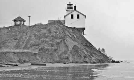 A small white house with a light tower adjacent sits on the edge of a cliff which has eroded away beneath it.