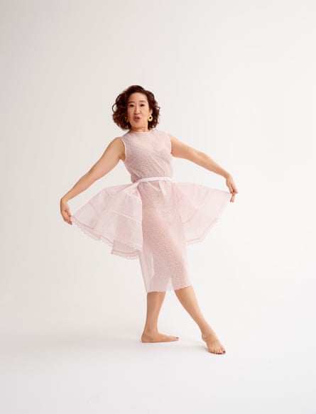 Oh wears a pink sleeveless organza midi dress and is barefoot, holding the side of the dress out a little like a tutu