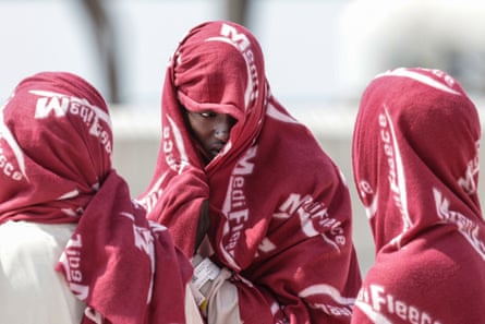 Three women wear red blankets over their heads and shoulders