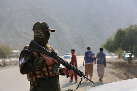 A Taliban fighter holding a firearm as three people walk away in the distance.