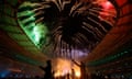 Silhouettes of dancers inside the Stade de France beneath red and green fireworks erupting across the stadium’s open roof