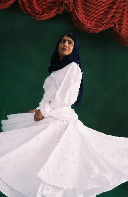 Malala Yousafzai wearing a long flowing white outfit and a black headscarf, standing against a dark-green wall