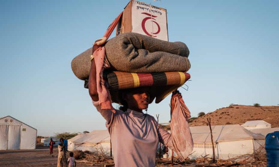A woman at Um Raquba refugee camp in Gedaref, eastern Sudan, balancing matting, a blanket and a box on her head as she walks past white tents