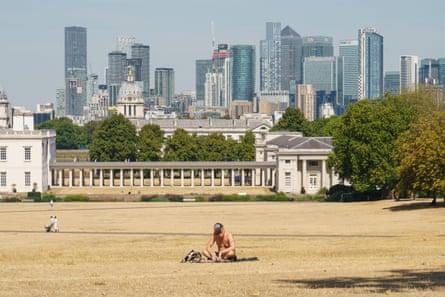 A man sits on the dried grass of Greenwich Park with skyscrapers in the background