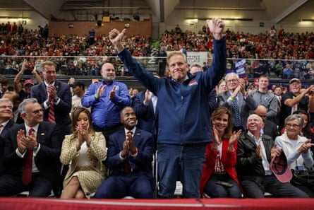 Man in navy blue sweater and jeans smiles and raises arms while standing as crowd claps