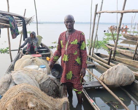 ‘The water is no longer our friend’: how dredging is pushing Lagos Lagoon towards ecosystem collapse – photo essay A man holding a bucket with a few fish in it stands next to a boat filled with fishing nets by the water.