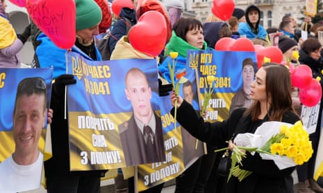 A woman in black hands out yellow flowers to the family members of Ukrainian prisoners of war, who hold red heart balloons and blue-and-yellow banners with their loved ones' faces on them. 