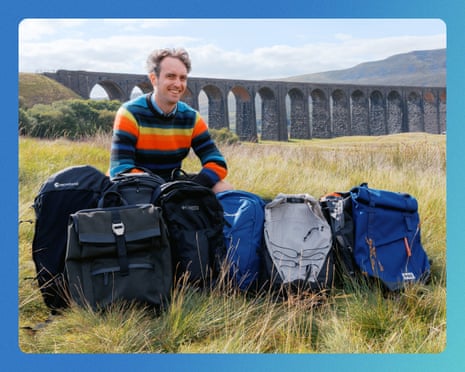 Pete Wise kneels behind a row of test backpacks in a field near a stone viaduct.
