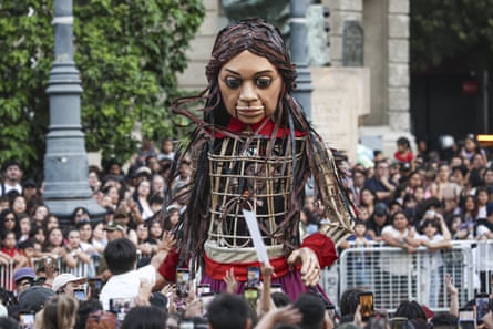 Lifesize herd of puppet animals begins climate action journey from Africa to Arctic Circle A large crowd of people watch a giant puppet of a girl on a street